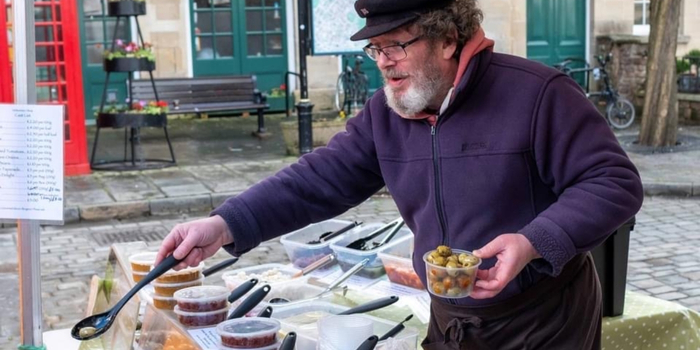 Image shows Richard Bardsley, Owner of Midsummer Olives serving green olives on a large black serving spoon at a market